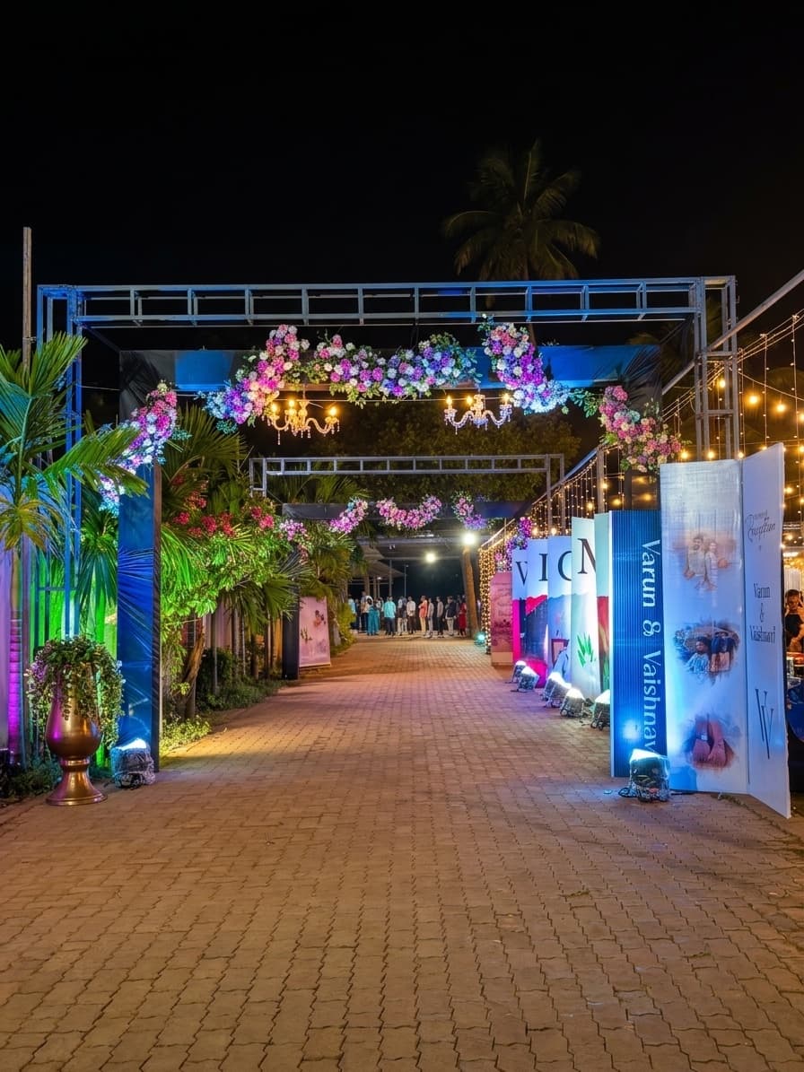 The entrance walk dressed for a wedding at night: a lit steel arch hung with pink and white flowers and two chandeliers, string lights strung through the palms, printed welcome boards along one side, and the brick paver walk lit warm underfoot.