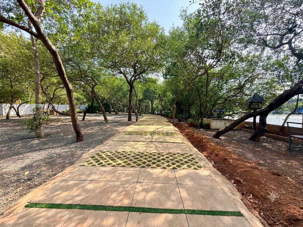 A paved walk through the grounds under a canopy of trees, with grass set in a chequered pattern between the tiles, gravel on either side, a bench and a bird feeder on the right, and the river showing beyond a low wall.