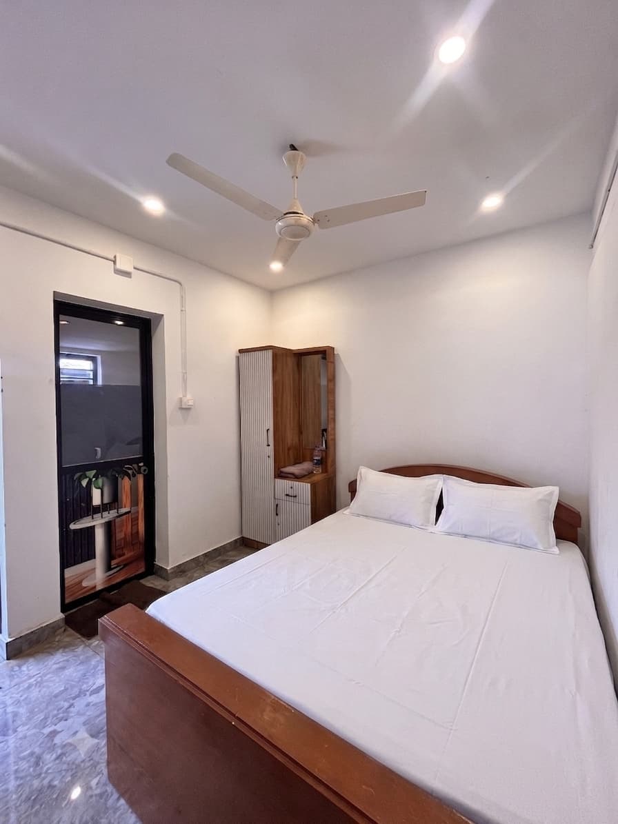 The fan-cooled Coconut Groove cottage: a double bed with a curved wooden headboard, a ceiling fan, a wardrobe with a mirror, and a glass door through to the bathroom.