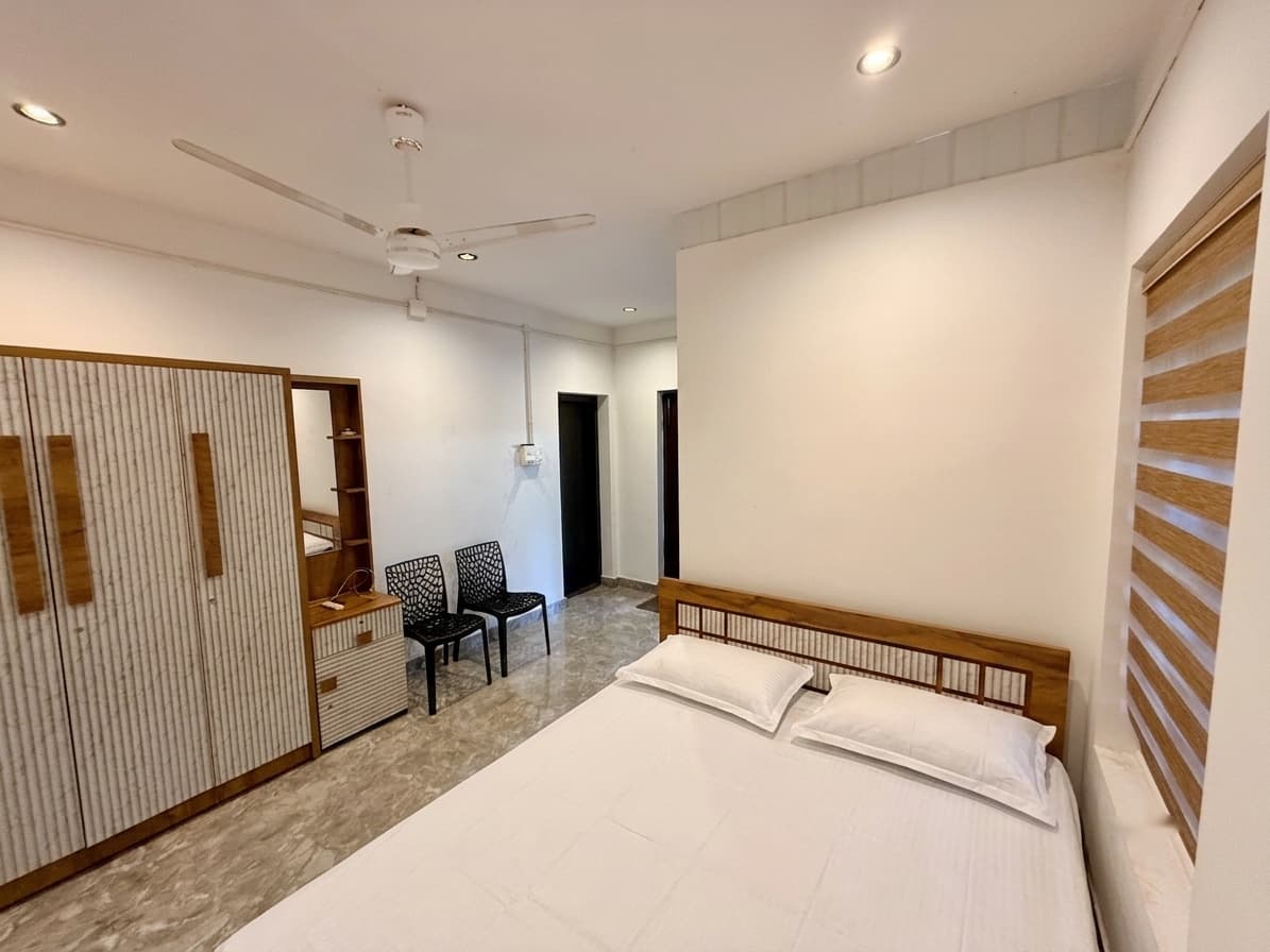 A Coconut Groove cottage room: a double bed with a slatted wooden headboard, a tall fluted wardrobe with a mirror, two chairs and a marble floor.