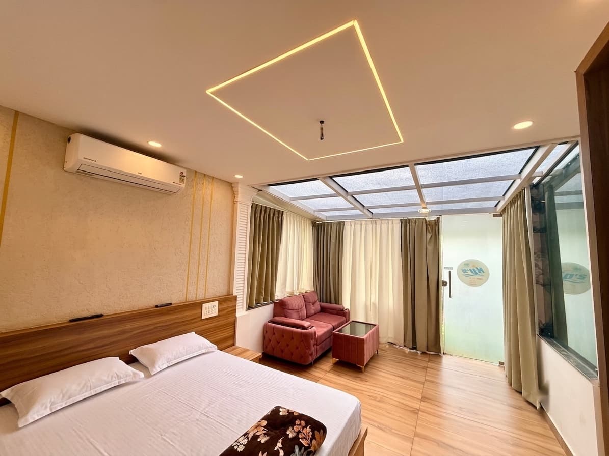 A River Breeze room: a wide bed against a wooden headboard, a red sofa and low table by the window, a split air conditioner and a recessed cove light, with a glass door onto the balcony.