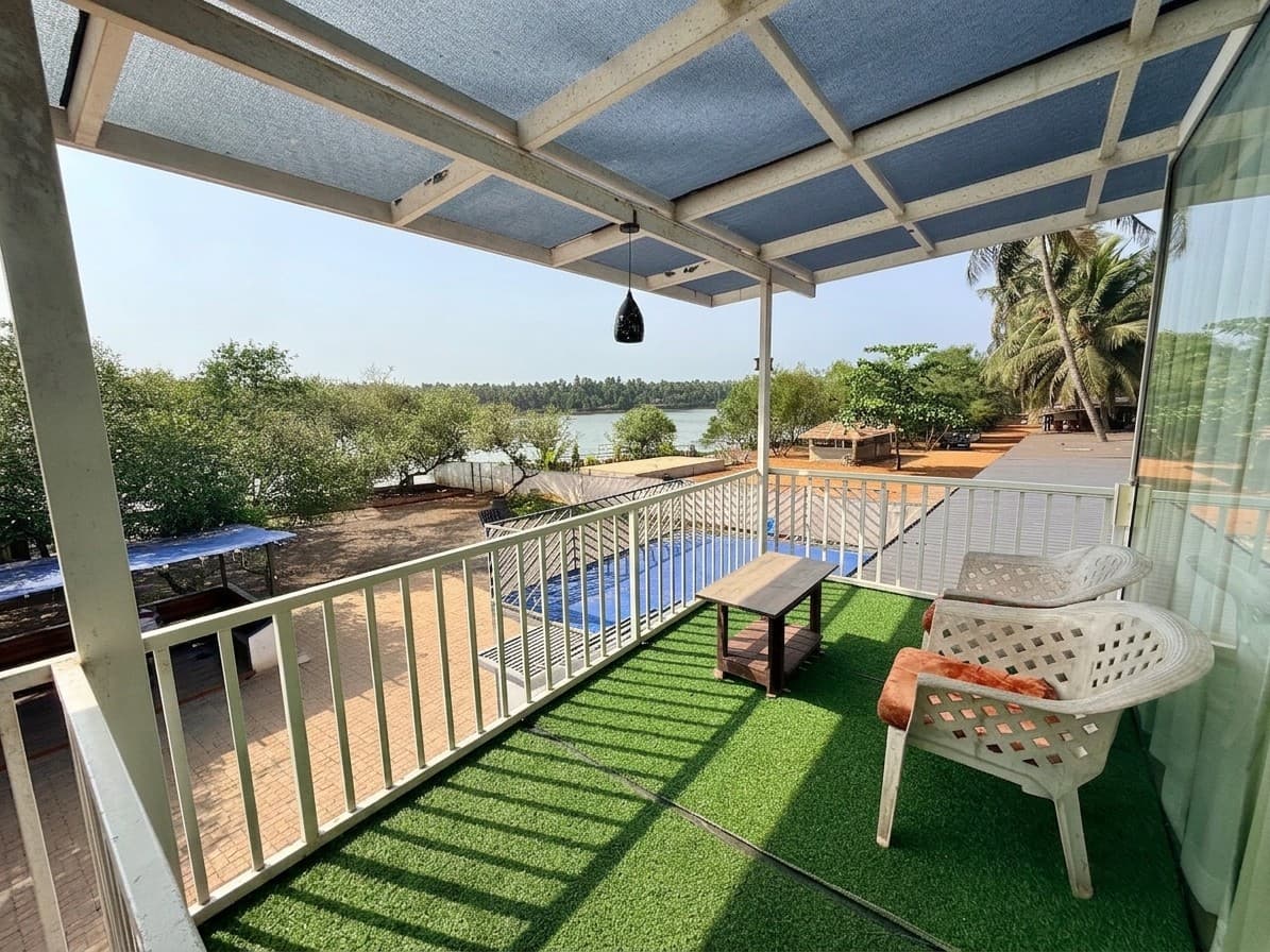 A River Breeze balcony under a slatted canopy: artificial grass, two white lattice chairs and a low wooden table, looking down over the pool and the grounds to the river.